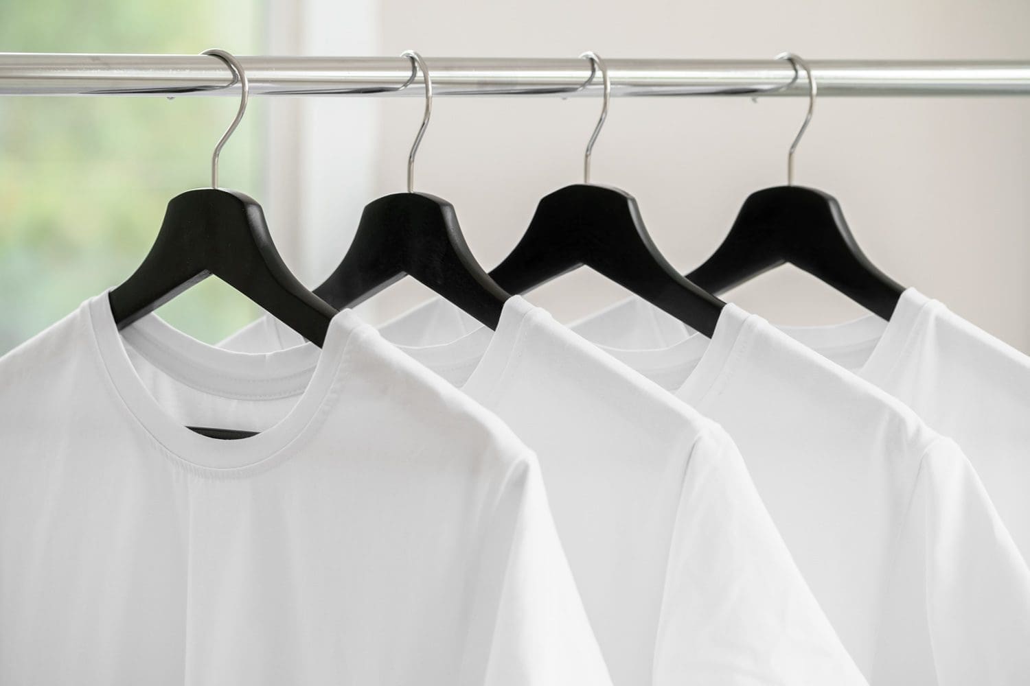 Four plain white t-shirts on black hangers are hanging on a metal clothes rack indoors.