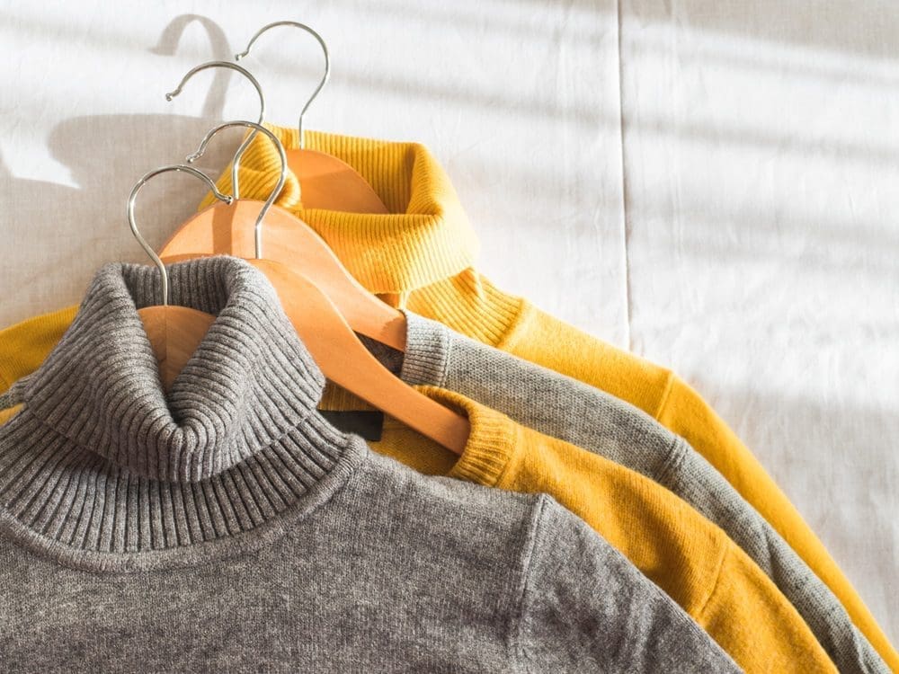 Three turtleneck sweaters, two yellow and one gray, are hung on wooden hangers against a light-colored background.