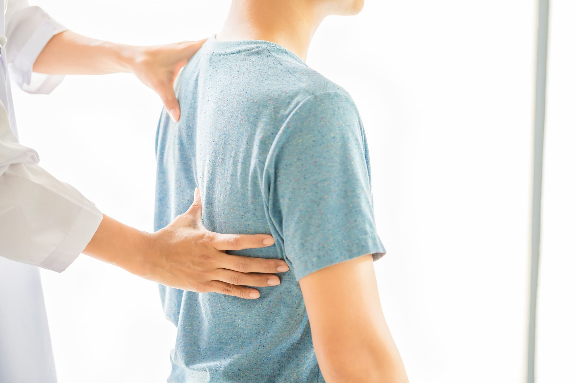 A person in a blue shirt receives a physical examination of the back by a healthcare professional.