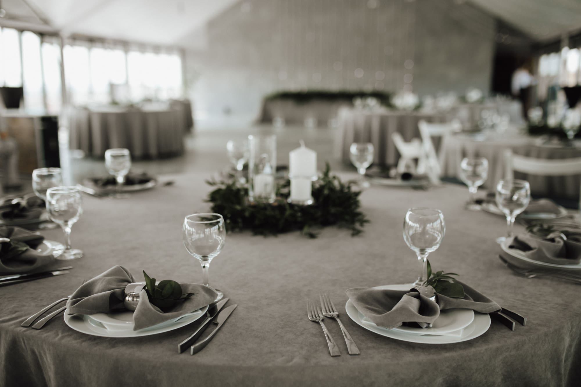 Round table set for a formal event with gray tablecloth, neatly folded napkins, glassware, cutlery, and a centerpiece of greenery and candles. Other tables visible in the background.