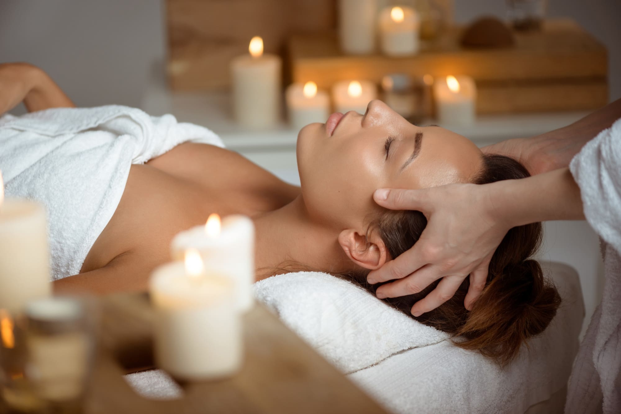 A person lies on a massage table with eyes closed, receiving a head massage surrounded by lit candles in a calm spa setting.