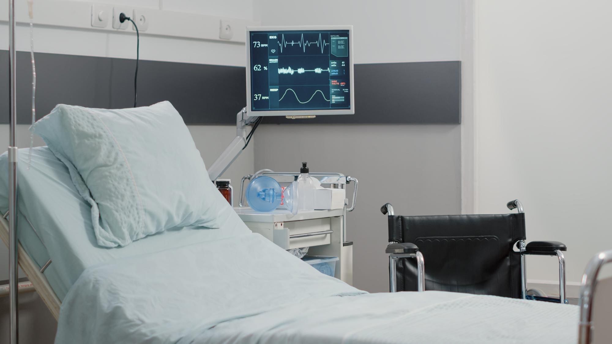 Hospital room with an empty bed, medical equipment, a monitor displaying vital signs, and a wheelchair beside the bed.