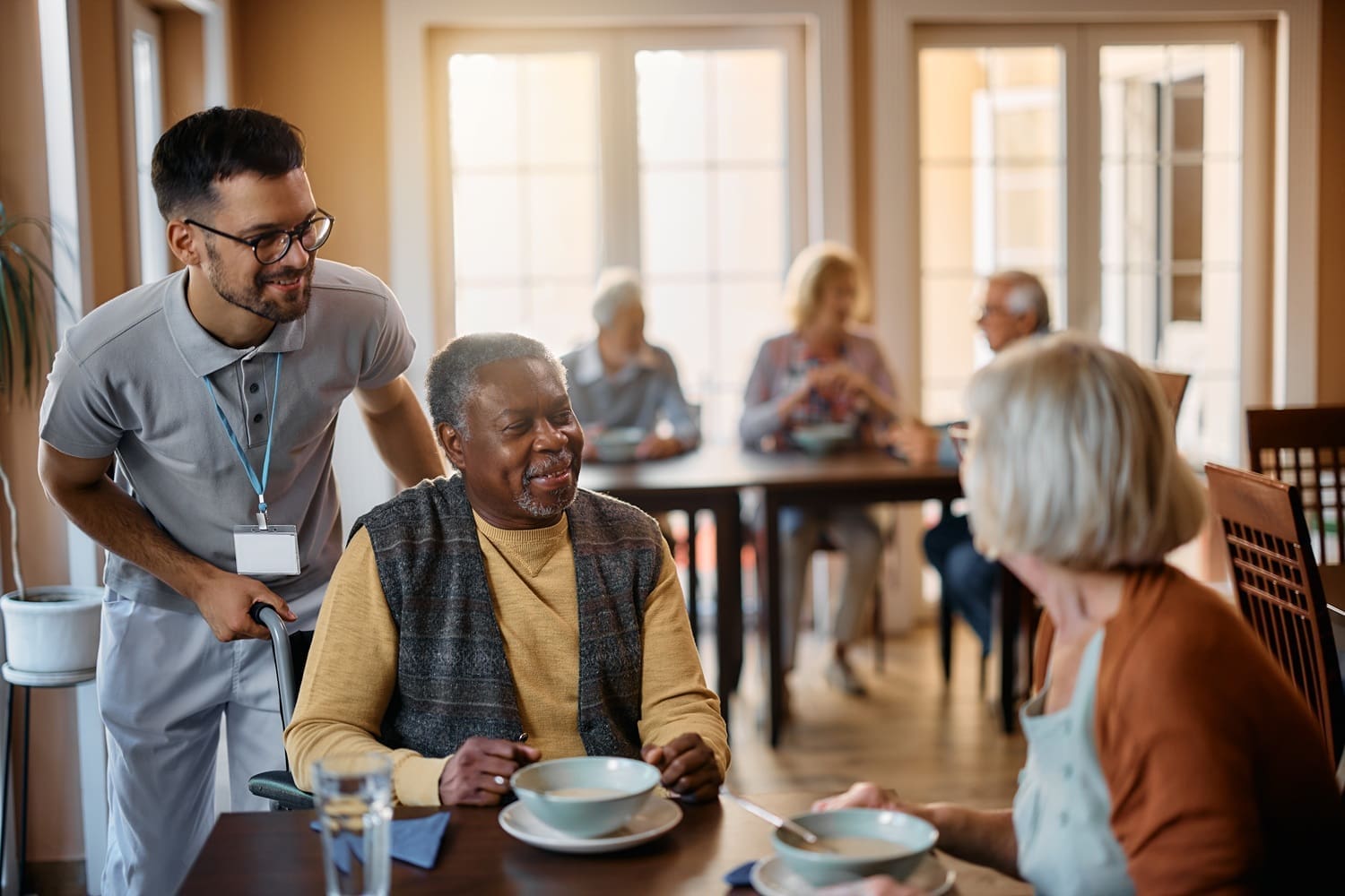 A caregiver assists an elderly man in a wheelchair while he talks with an elderly woman at a dining table in a communal setting; other seniors are in the background.