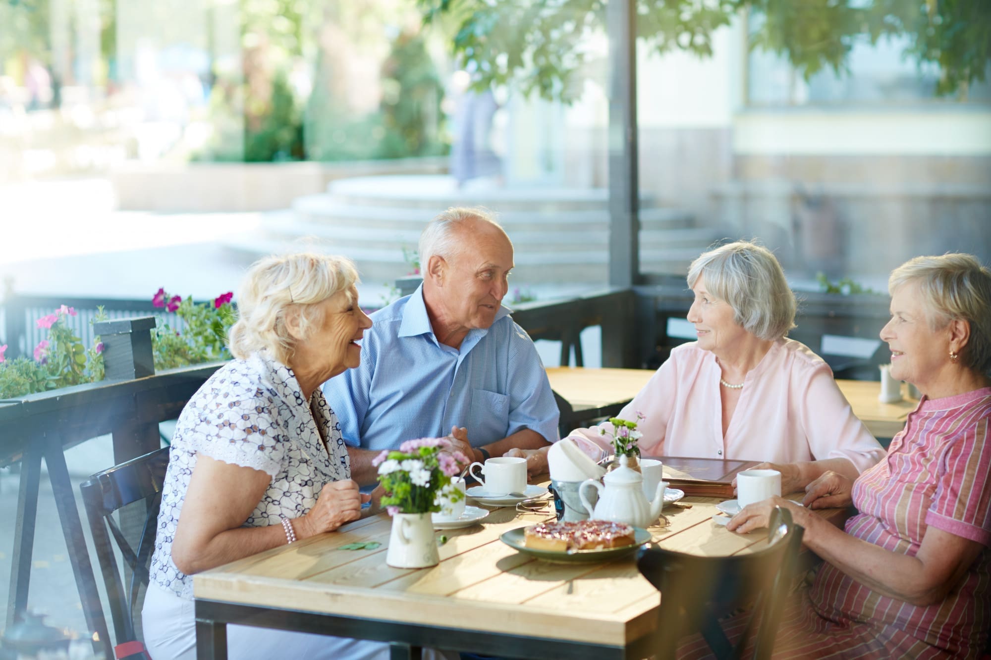 Four older adults sit at an outdoor café table, conversing and drinking coffee with pastries and flowers on the table, surrounded by greenery.