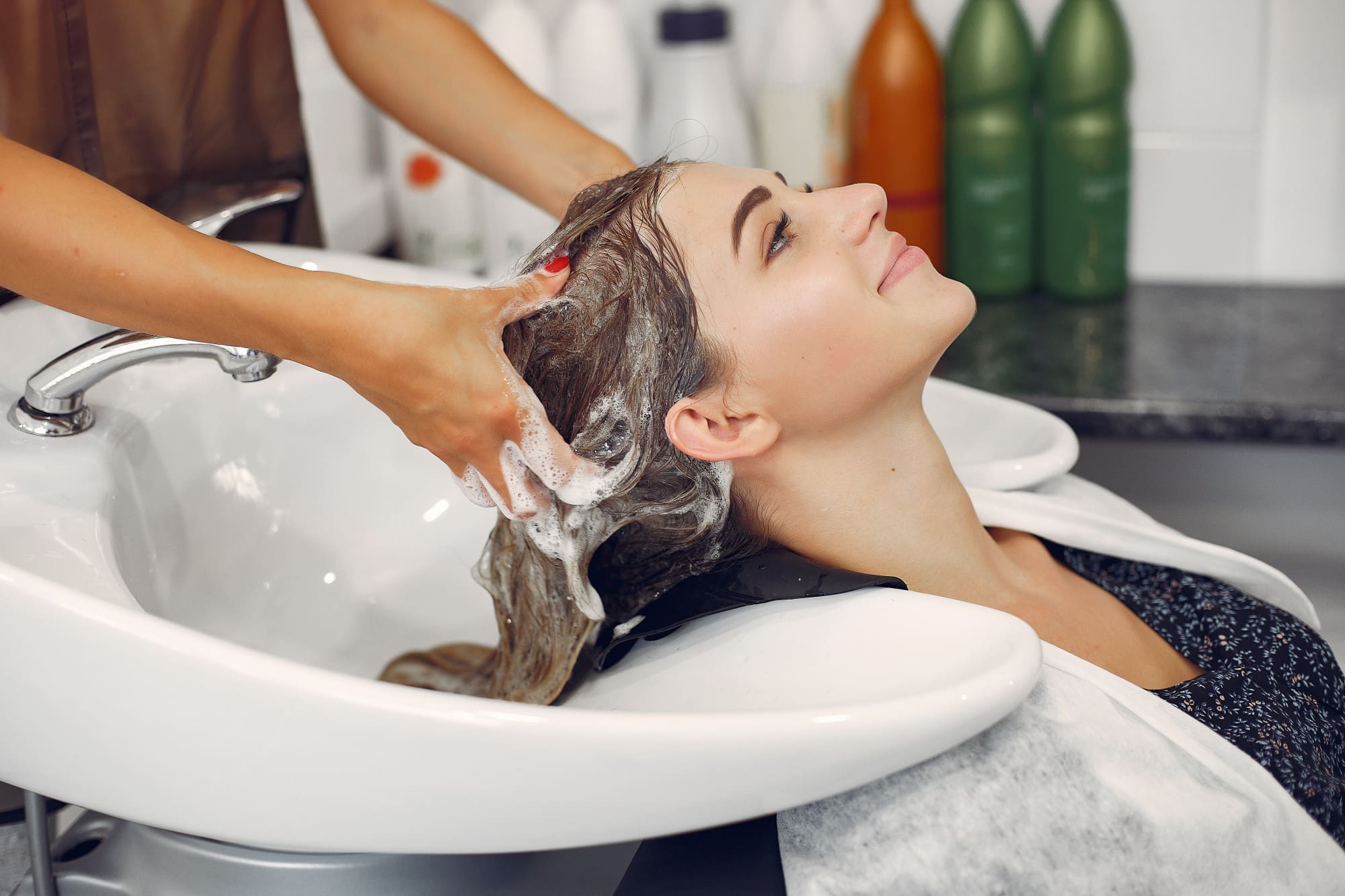 A woman is having her hair washed at a salon sink by a stylist, with shampoo lather visible in her hair.