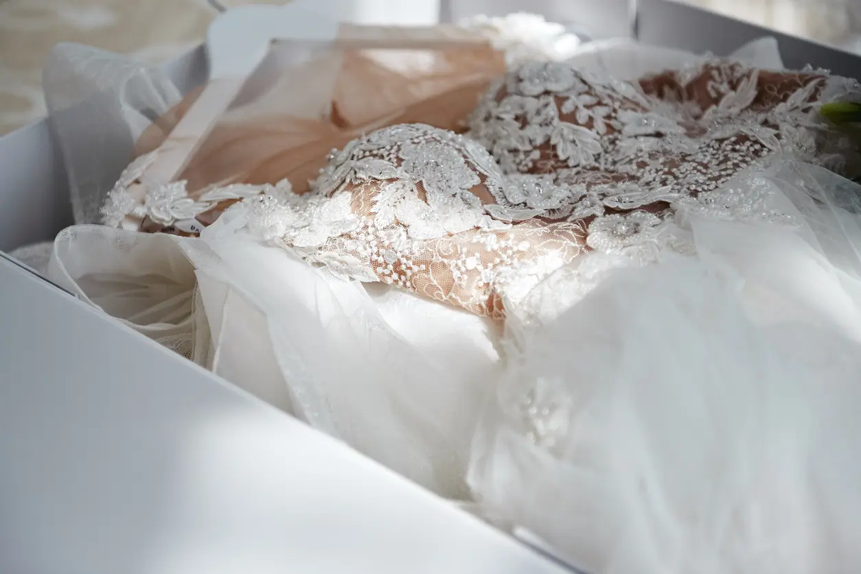 A close-up of a delicate white lace wedding dress with floral embroidery, neatly folded inside a white box.